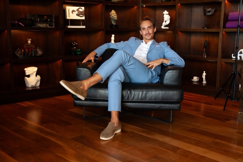 Benza Maman in a light blue suit, sitting in a black leather armchair in front of a lit bookshelf