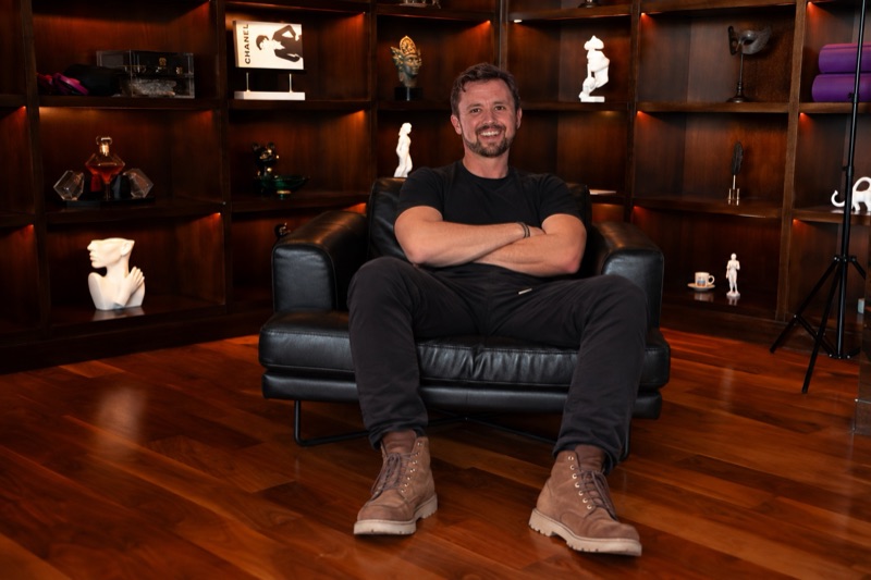 Chaz Bademan smiling with his arms crossed, sitting in a black leather armchair in front of lit wooden shelves